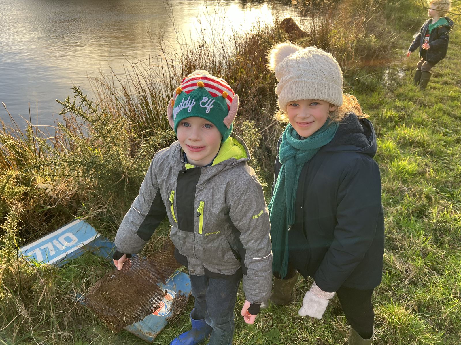Two children in winter coats and woolly hats on the grassy bank of the farm's fishing lake, with a third child behind them and low winter sun on the water.